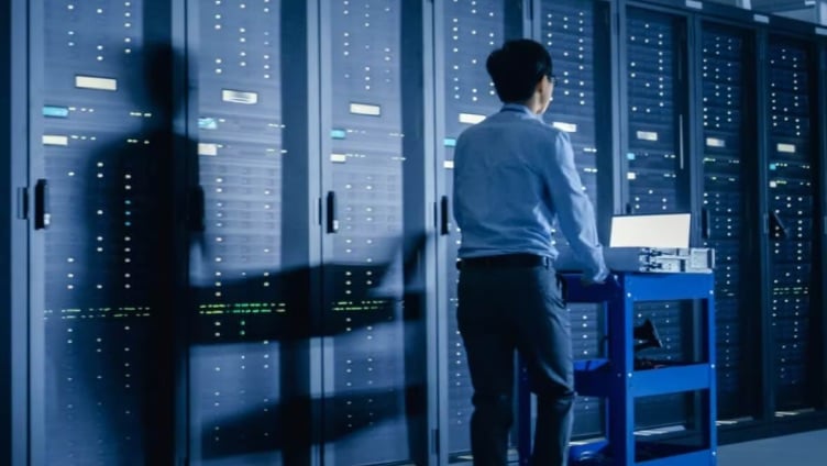 Une personne se tient dans l'allée d'un data center et travaille avec des équipements devant de grands racks de serveurs.