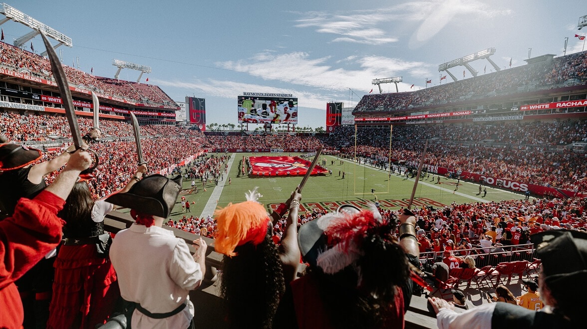 Crowd-filled football stadium during a pregame ceremony, viewed from the stands. Spectators in pirate-themed costumes raise swords in the foreground while a large team flag is displayed on the field beneath a bright sky.