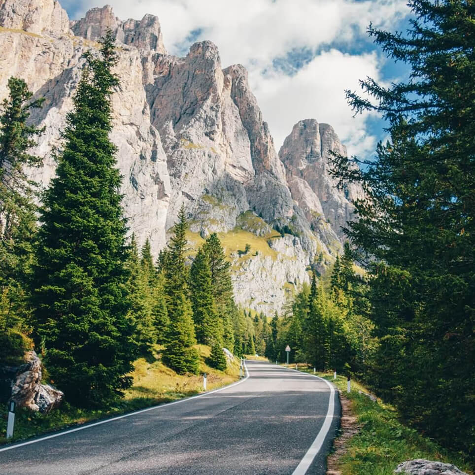 Scenic mountain road winding through a forest with rocky cliffs and evergreen trees under a bright blue sky