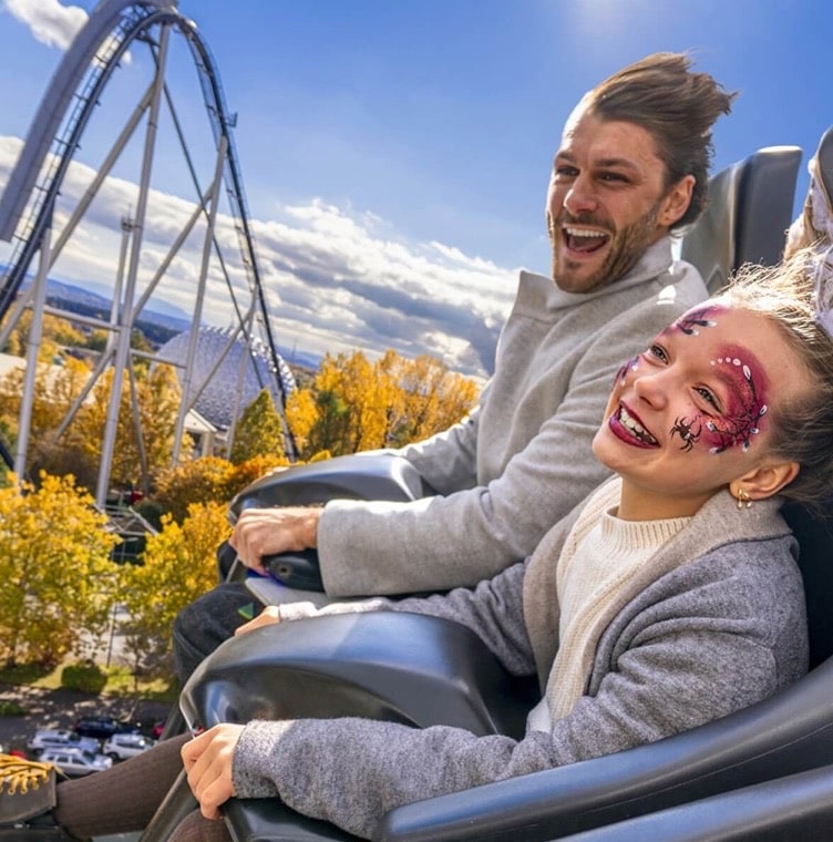 People riding a roller coaster at an amusement park with a large coaster track in the background.