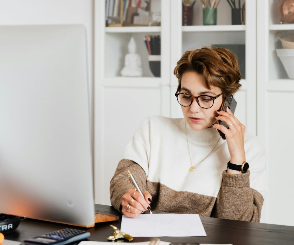 Person talking on a smartphone while writing notes at a desk in a modern home office setting.