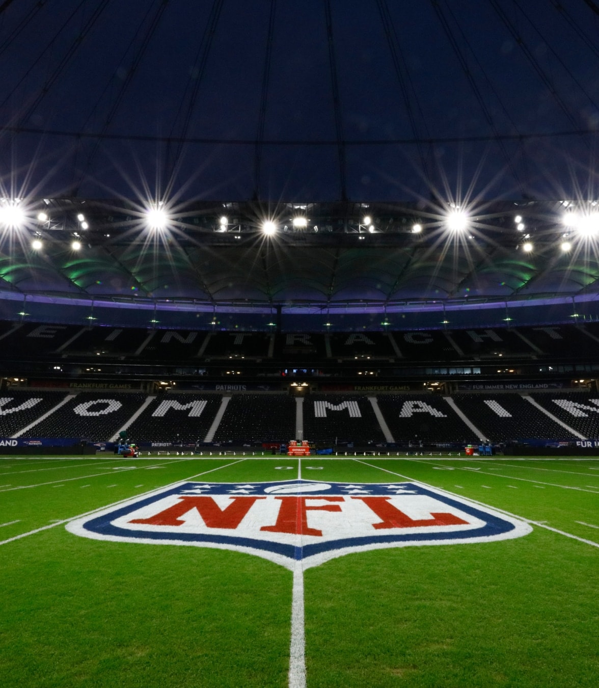 Vista nocturna de un campo de fútbol americano de la NFL iluminado con el logotipo de la NFL en el centro del campo dentro de un gran estadio.