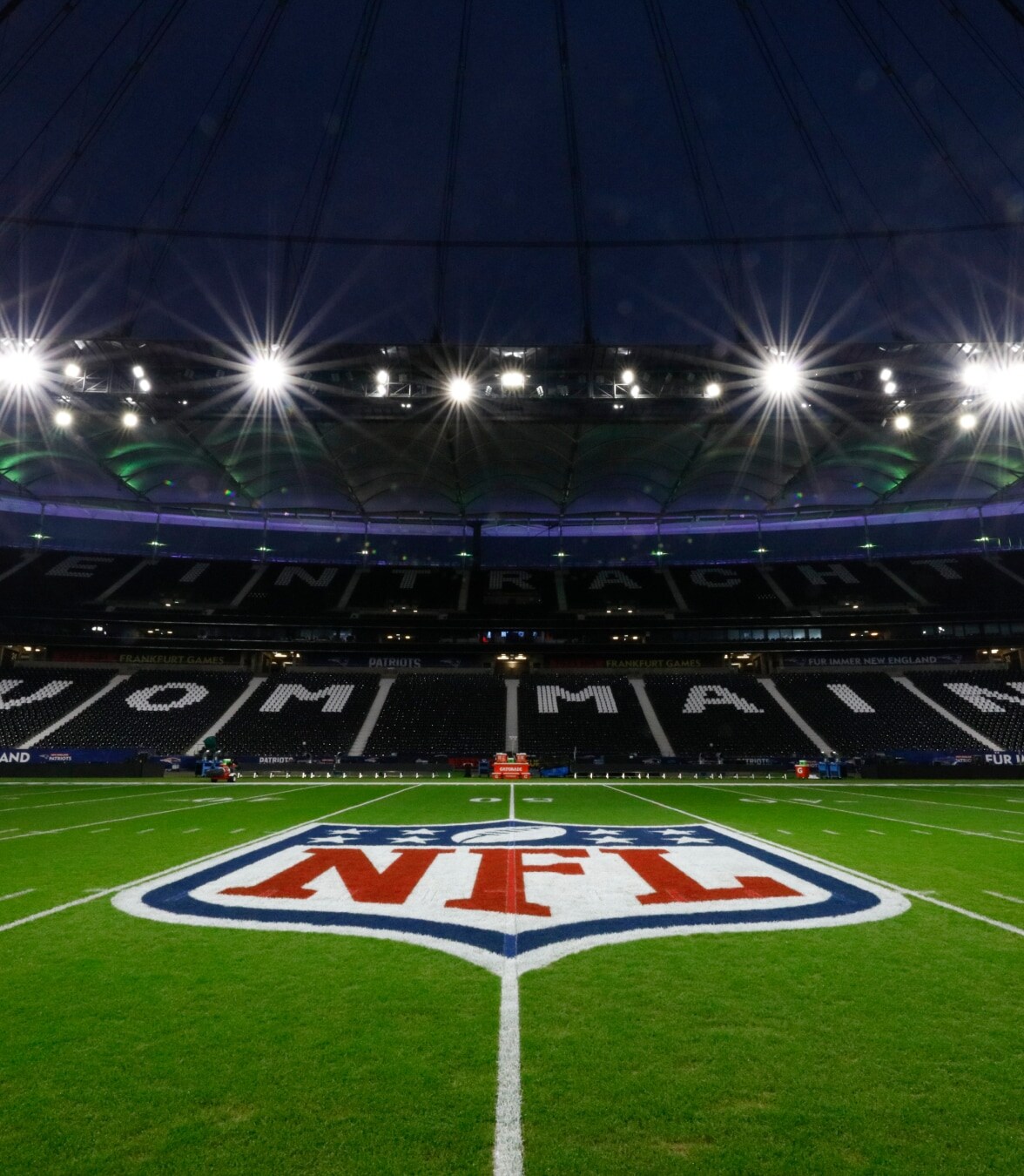 Vue nocturne d'un terrain de football américain illuminé avec le logo NFL au centre, à l'intérieur d'un grand stade.