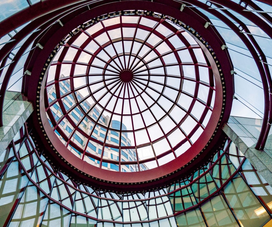Looking up through a circular glass atrium roof with a red steel framework, surrounded by modern office building windows.