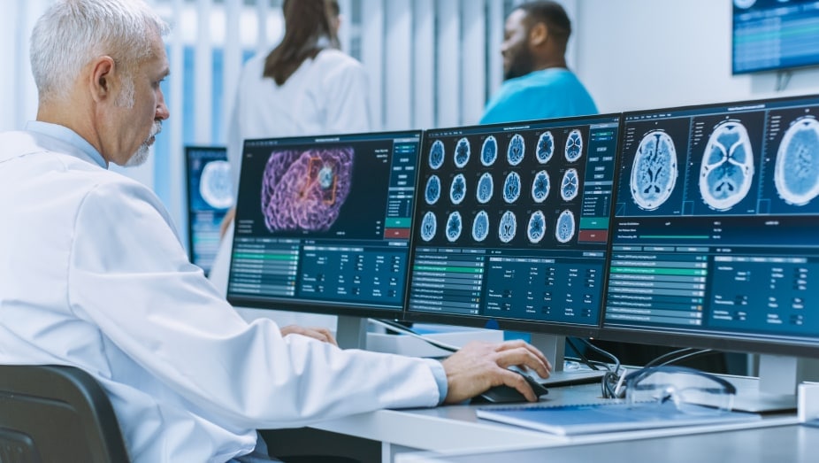 A medical professional sits facing several computer screens displaying medical imaging data.