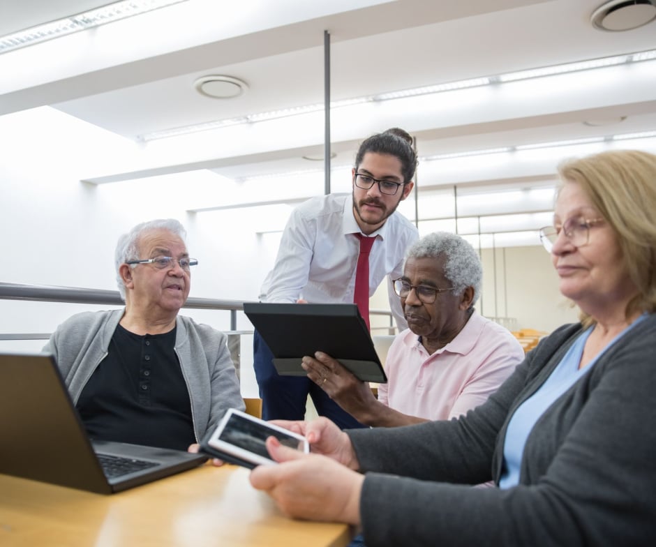 People in a learning session use a laptop, tablet, and smartphone while receiving digital skills guidance in a bright classroom setting.