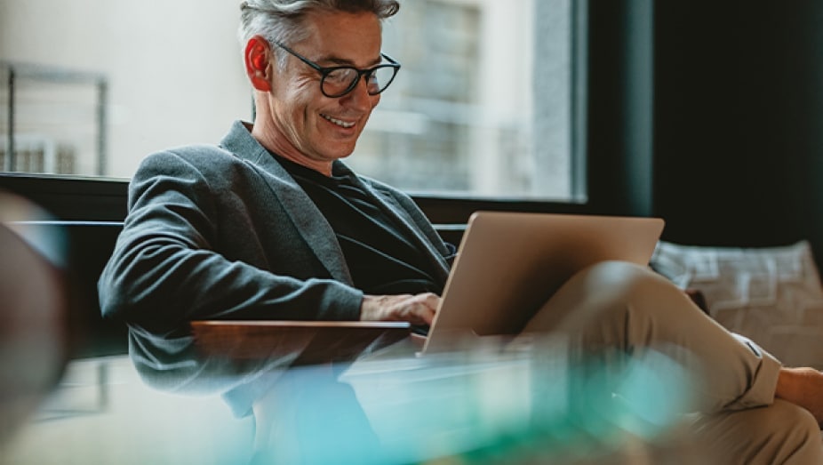 A stylish man sits in a lounge chair and smiles while looking at an open laptop.