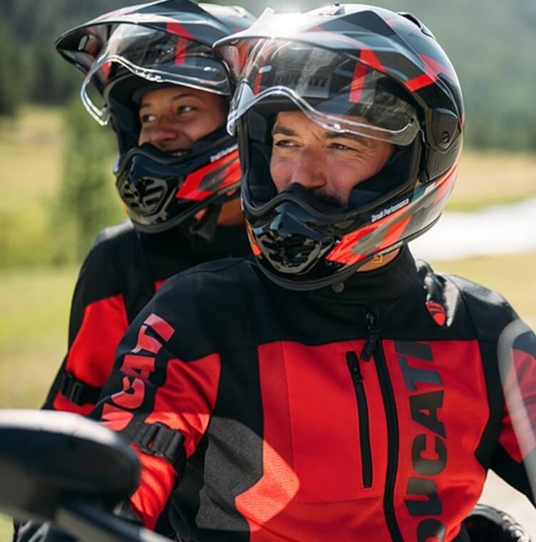 A couple seated on a Ducati motorcycle, wearing Ducati touring apparel in a mountainous setting