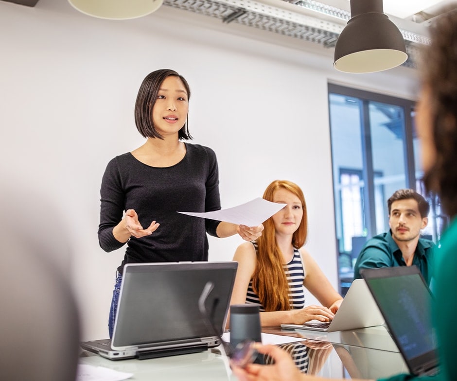 Lady holding a paper and speaking in front of her laptop