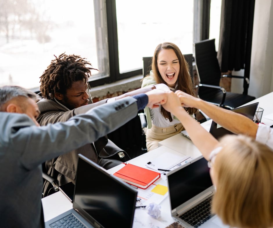 Team members gathered around a conference table, joining hands in the center during a collaborative office meeting.
