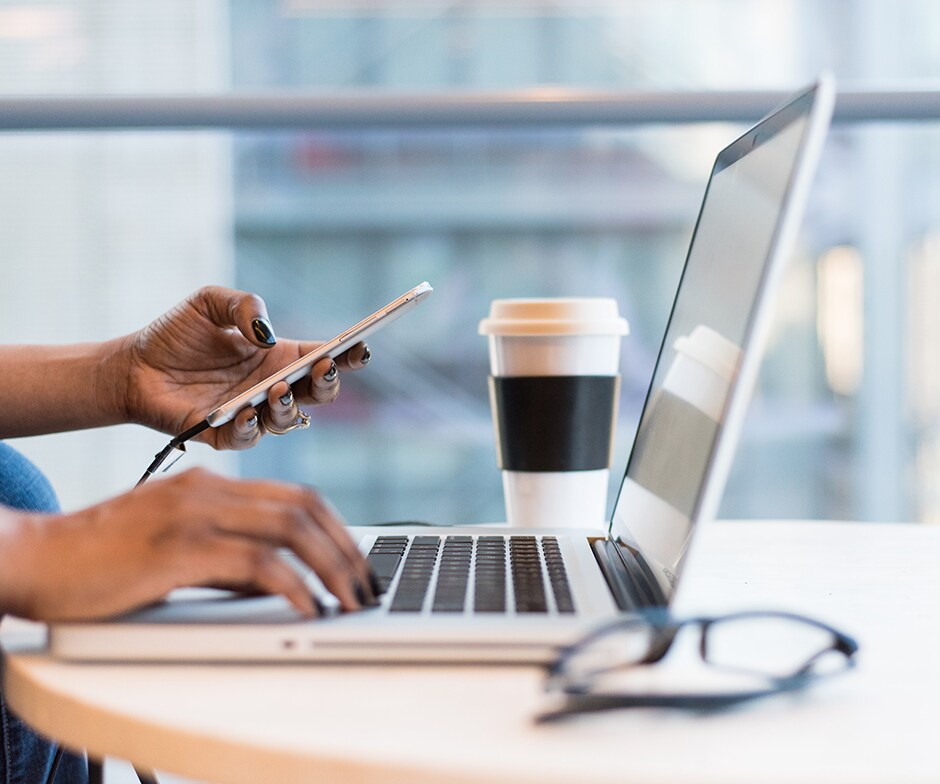 vista ampliada de una estación de trabajo con un portátil, gafas para el ordenador, una taza de café y las manos de una persona (una en el teclado y la otra sosteniendo un smartphone)